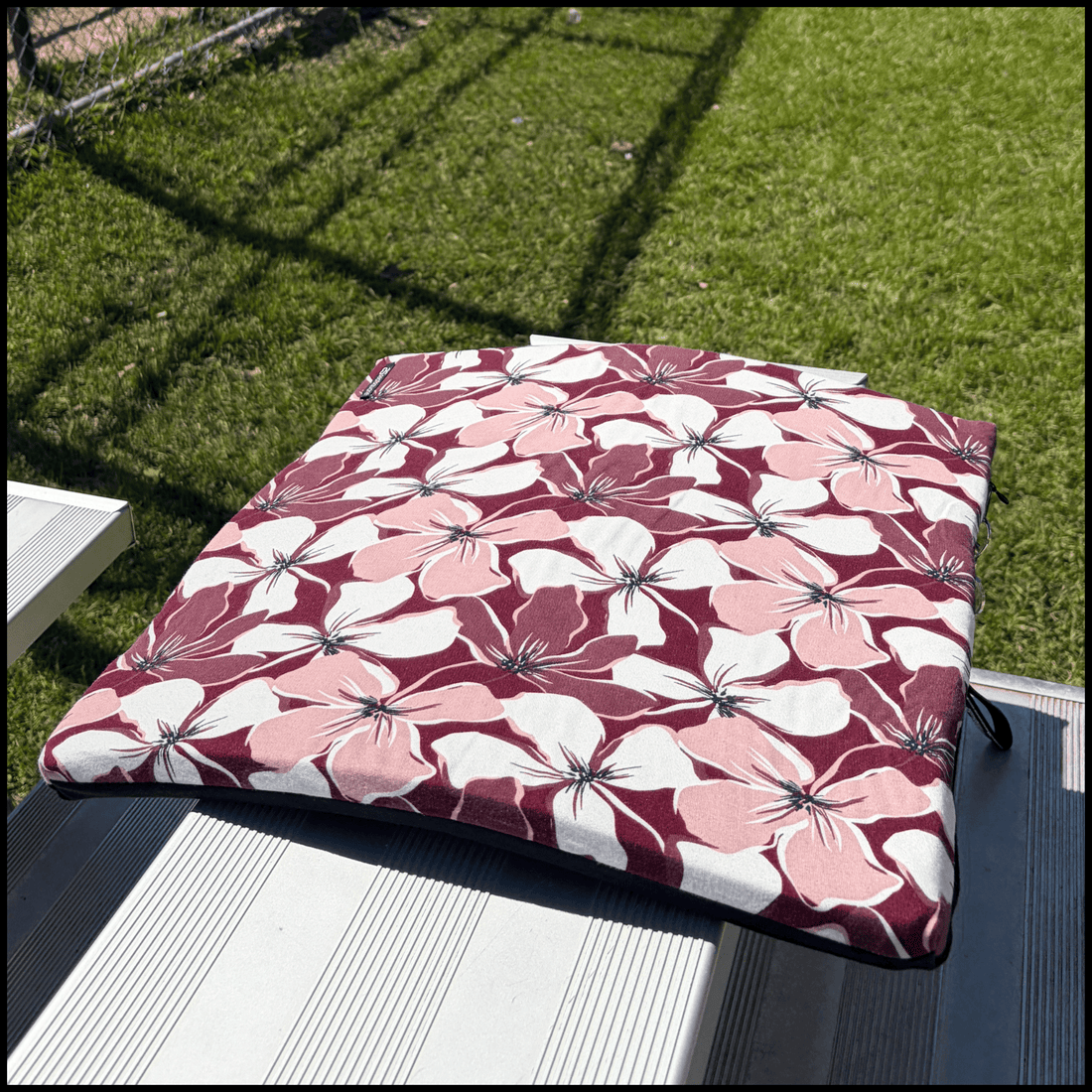A beautiful square seat cushion with a bold floral pattern in burgundy, light pink, and white, on a metal bleacher at a baseball diamond 