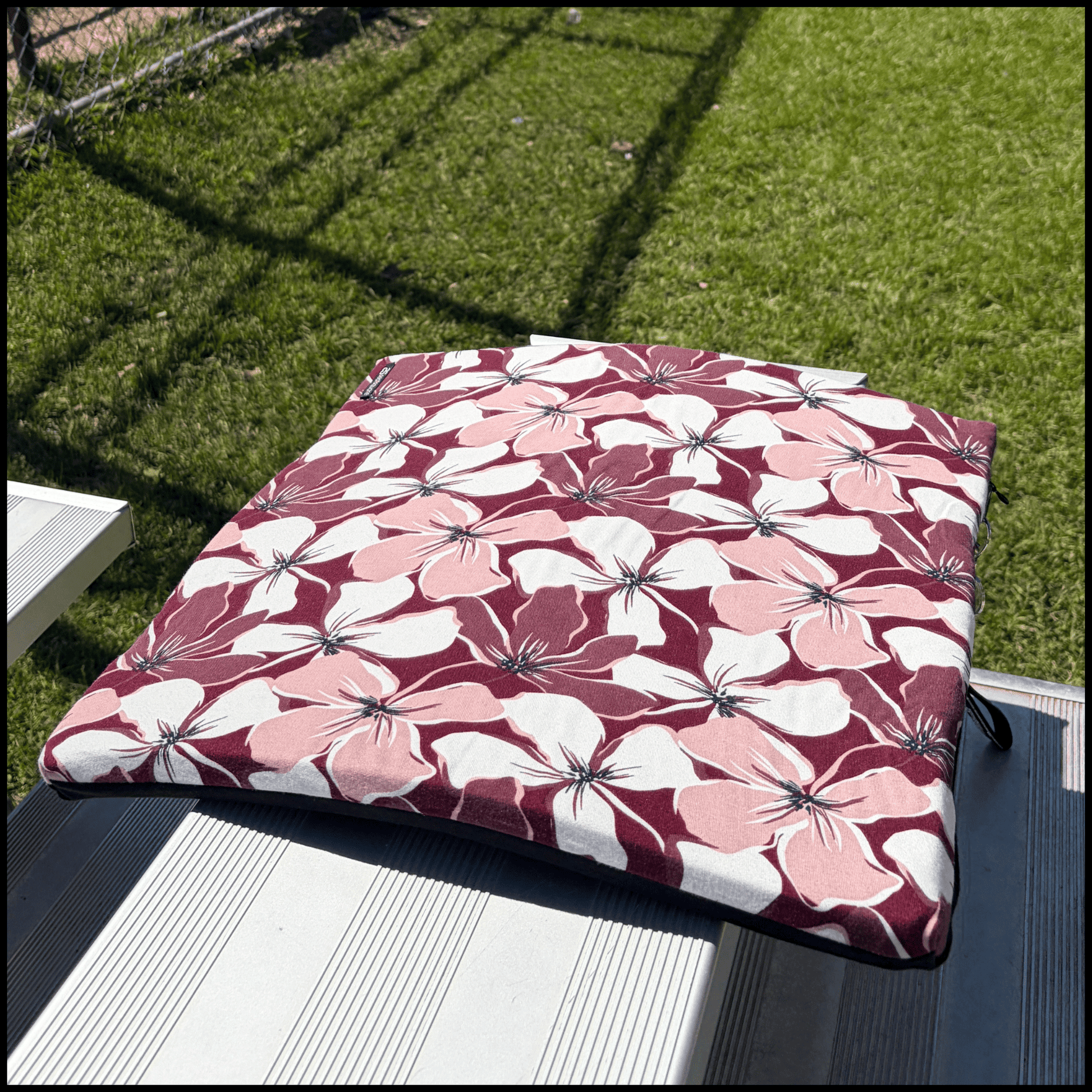 A beautiful square seat cushion with a bold floral pattern in burgundy, light pink, and white, on a metal bleacher at a baseball diamond 
